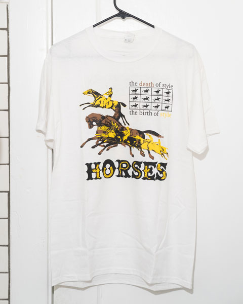 a white  graphic t-shirt on a hanger. The graphic is printed in yellow, brown and black. it shows a group of horses in a flying gallop with a Muybridge sequence above that. Below everything written in a western font is the word horses