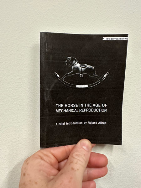 a hand holding up a small zine printed in black ink. The title of the zine is The Horse in the Age of Mechanical Reproduction